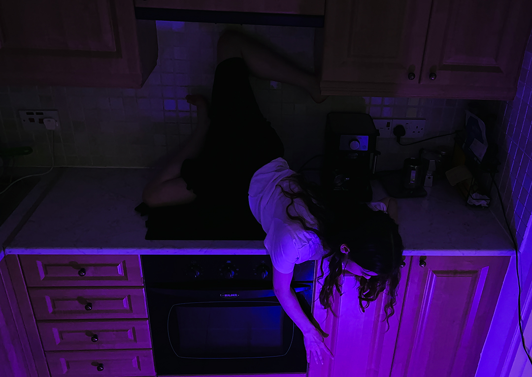 Dancing in my underpants. Picture from the photobook. Me on my kitchen stove reaching down towards a purple light, the room is dark.